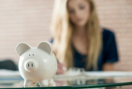 A high school teenager is writing down monthly budget in her notebook. A piggy bank is sitting on the table.